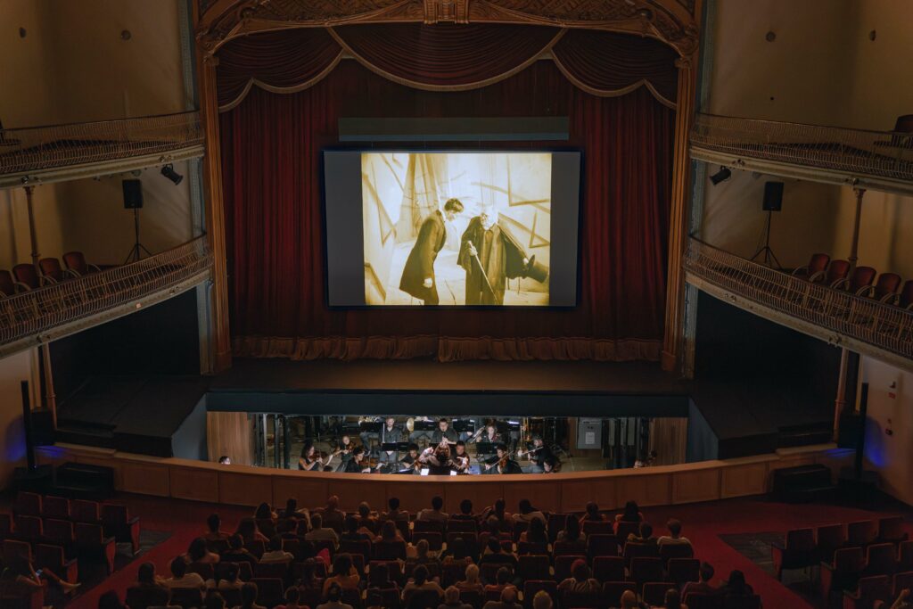 PraTodosVerem: Foto de uma sala de espetáculos com um telão exibindo um filme em preto e branco com legendas em português. Na parte inferior, é possível ver a orquestra tocando no fosso, enquanto o público assiste à projeção nas poltronas do teatro. O espaço possui um palco ornamentado, cortinas vermelhas e dois andares de balcões. Foto: Íris Zanetti